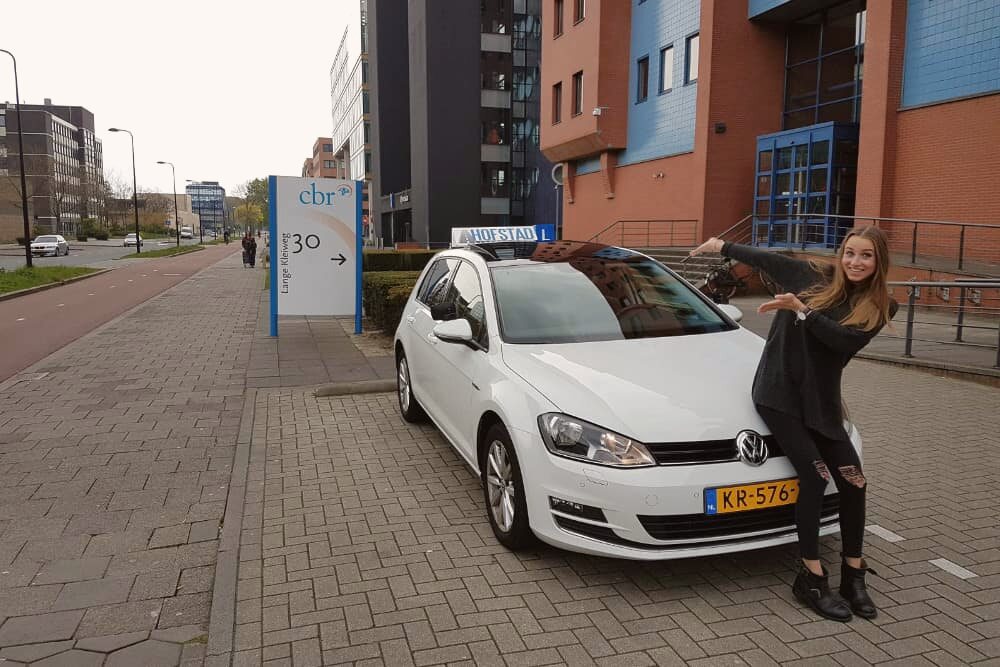 Geslaagde leerling wijst trots naar de HOFSTAD-daklichtbak op de witte lesauto, bij het CBR in Rijswijk